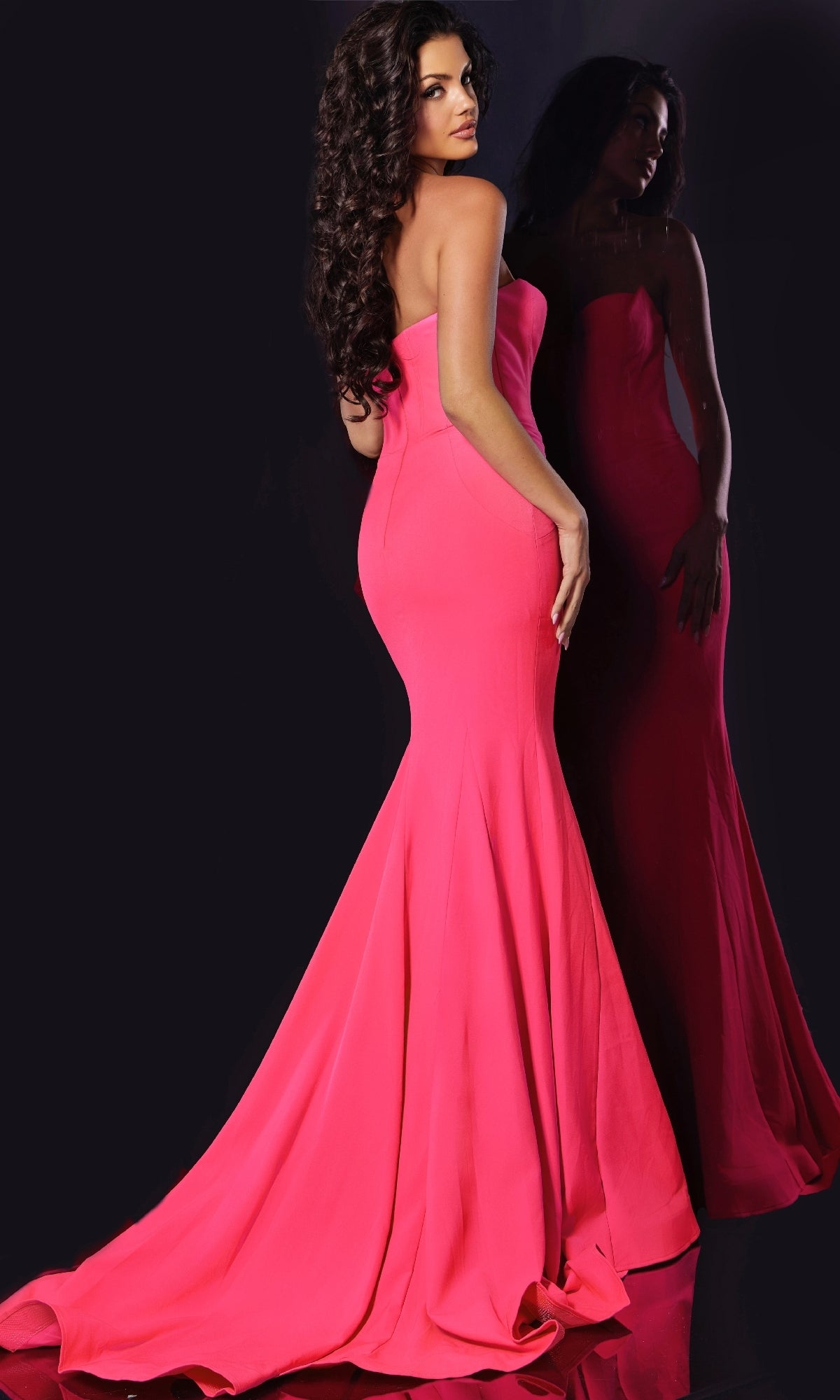 A woman stands before a mirror in the Jovani 38974 long prom dress. The fitted, strapless pink gown reflects her elegance and poise against a dark backdrop.